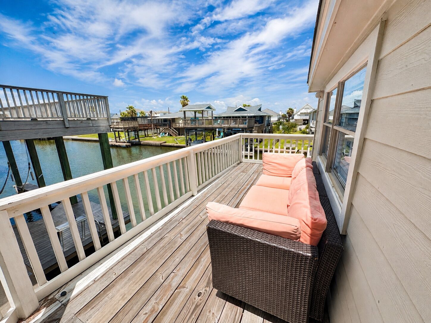 A wooden deck with a wicker sofa and cushions, overlooking a marina with boats and a dock.