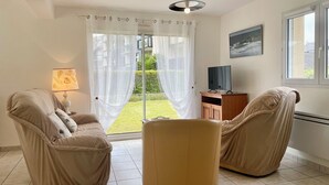 A room with two beige armchairs, a small table, a TV on a wooden stand, and a large window with a view of a garden. - L'Irisée-6p-Quiberon-500m Thalasso and beach parking lot (Quiberon)