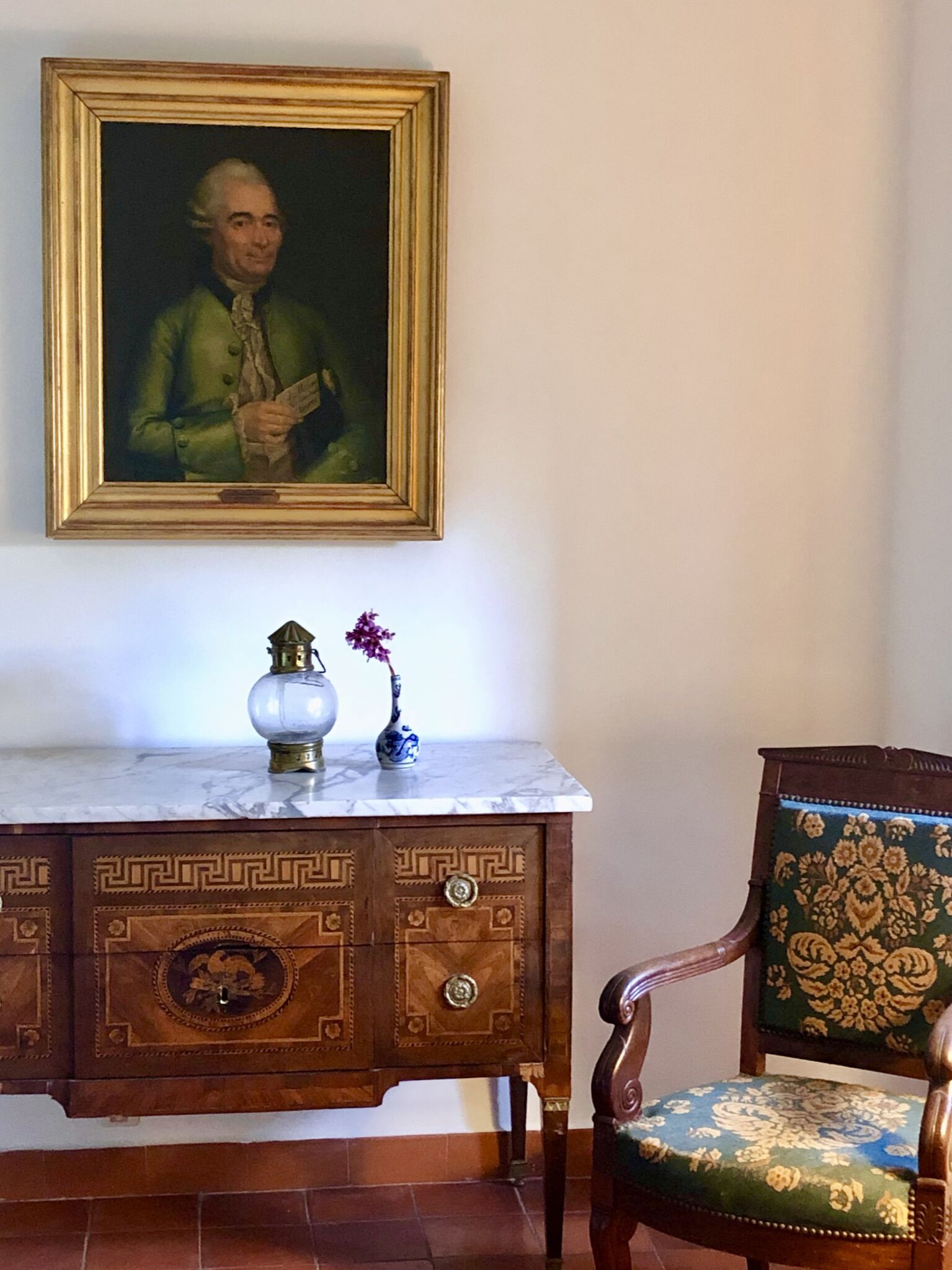 Una habitación con un escritorio de encimera de mármol, un retrato enmarco, un florero con flores y una silla estampada.