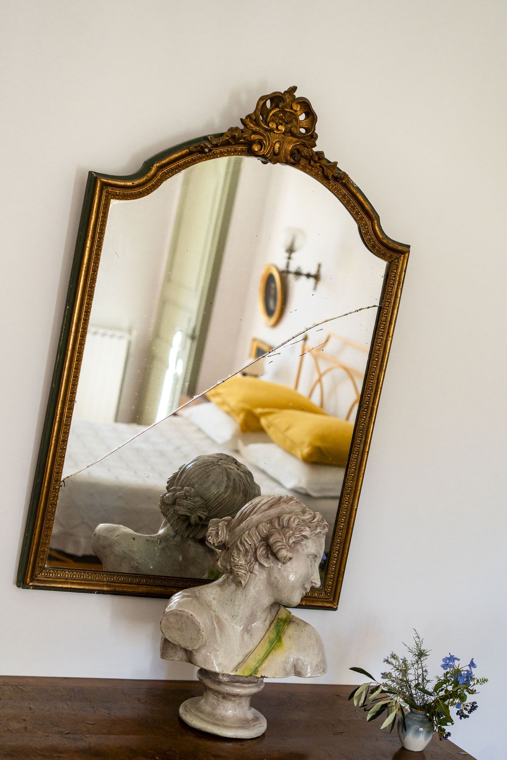 A mirror with an ornate frame reflecting a bed with yellow bedding and a statue on a wooden surface below.