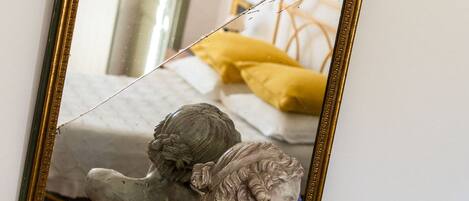 A mirror with an ornate frame reflecting a bed with yellow bedding and a statue on a wooden surface below.