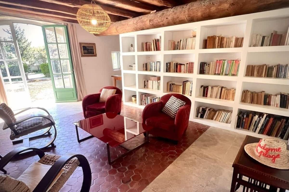 A cozy living room with a glass coffee table, two red armchairs, a bookshelf filled with books, and a wooden dining table with a cake.