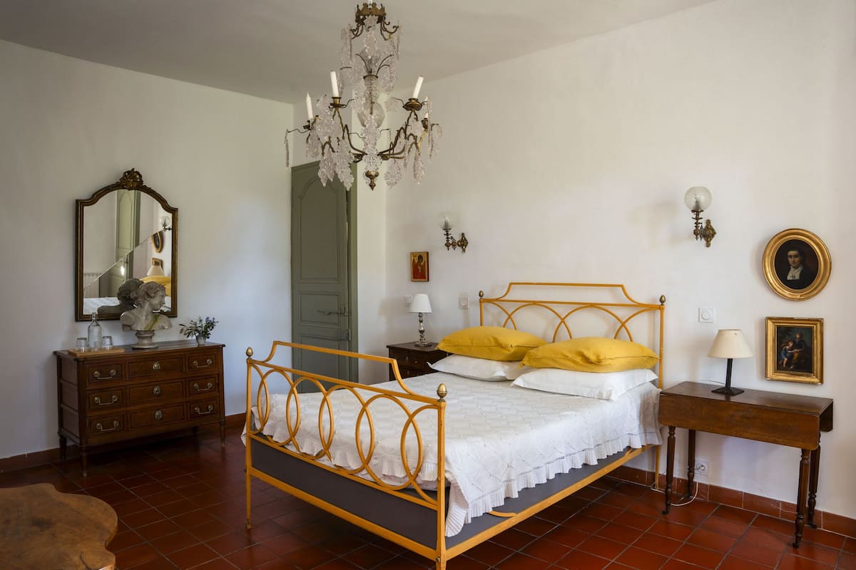 A bedroom with a wooden four-poster bed, a chandelier, a dresser, a mirror, and a bedside table with a lamp.