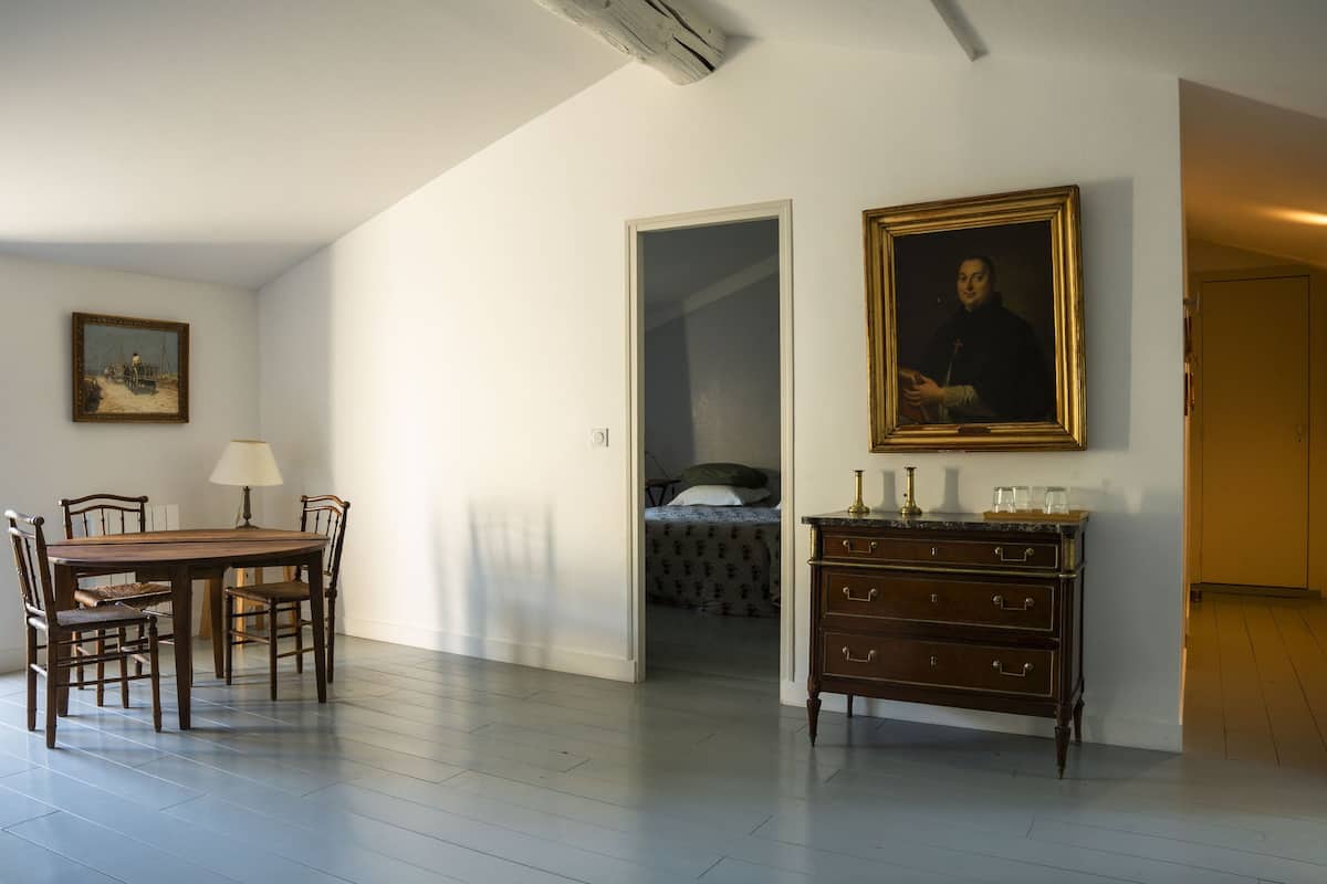 A dining area with a wooden table and chairs, a dresser with drawers, a framed portrait on the wall, and a bed in the adjacent room.