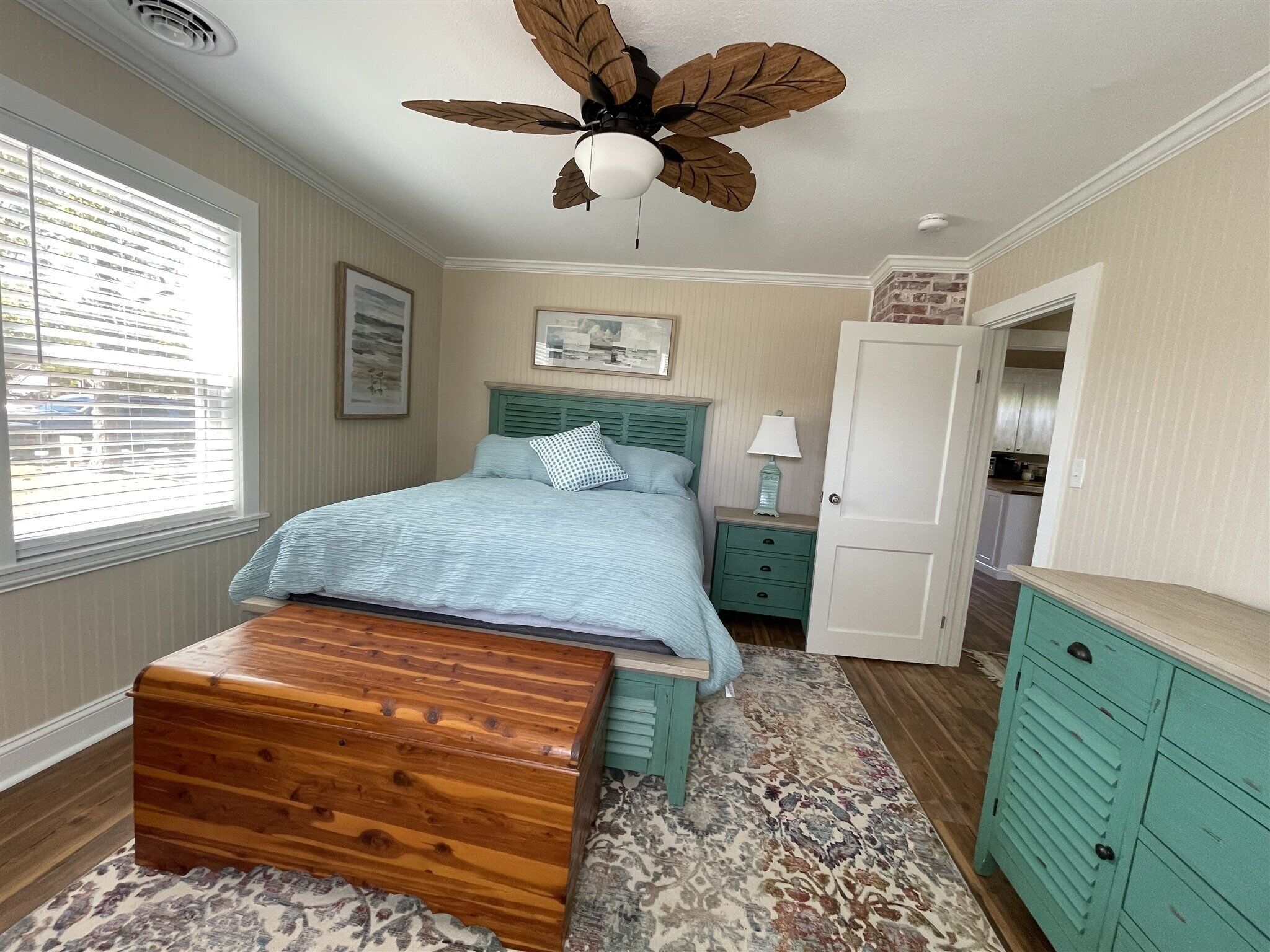 Une chambre à coucher avec un lit, une commode en bois, une table de chevet, une lampe, une porte et une vue sur l’extérieur par une fenêtre.
