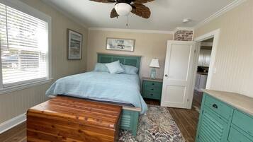 Une chambre à coucher avec un lit, une commode en bois, une table de chevet, une lampe, une porte et une vue sur l’extérieur par une fenêtre.