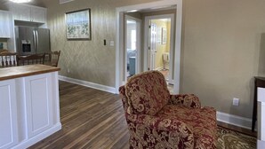A spacious living area with a wooden floor, a patterned armchair, and a kitchen with white cabinetry and a stainless steel refrigerator. - Gracie's Place 2 Bedroom Cottage (Avon)