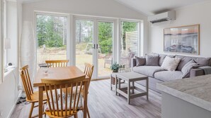 Una sala de estar moderna con comedor, grandes puertas correderas de vidrio hacia un jardín, un sofá y un cuadro en la pared.