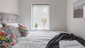 Un dormitorio con cama, ventana con vistas, mesita de noche con lámpara y un cuadro enmarcado en la pared.