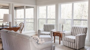 A bright, spacious room with large windows, a wooden desk, two striped armchairs, and a grey sofa.