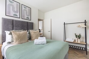 A neatly made bed with a green bedspread, two pillows, and a folded towel. There is a wall-mounted shelf with a plant and a hanging rack with clothes.