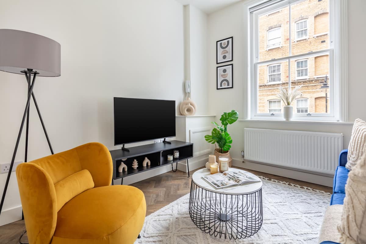 Een moderne woonkamer met een flatscreen televisie, een gele fauteuil, een ronde salontafel en een groot raam met uitzicht op gebouwen.
