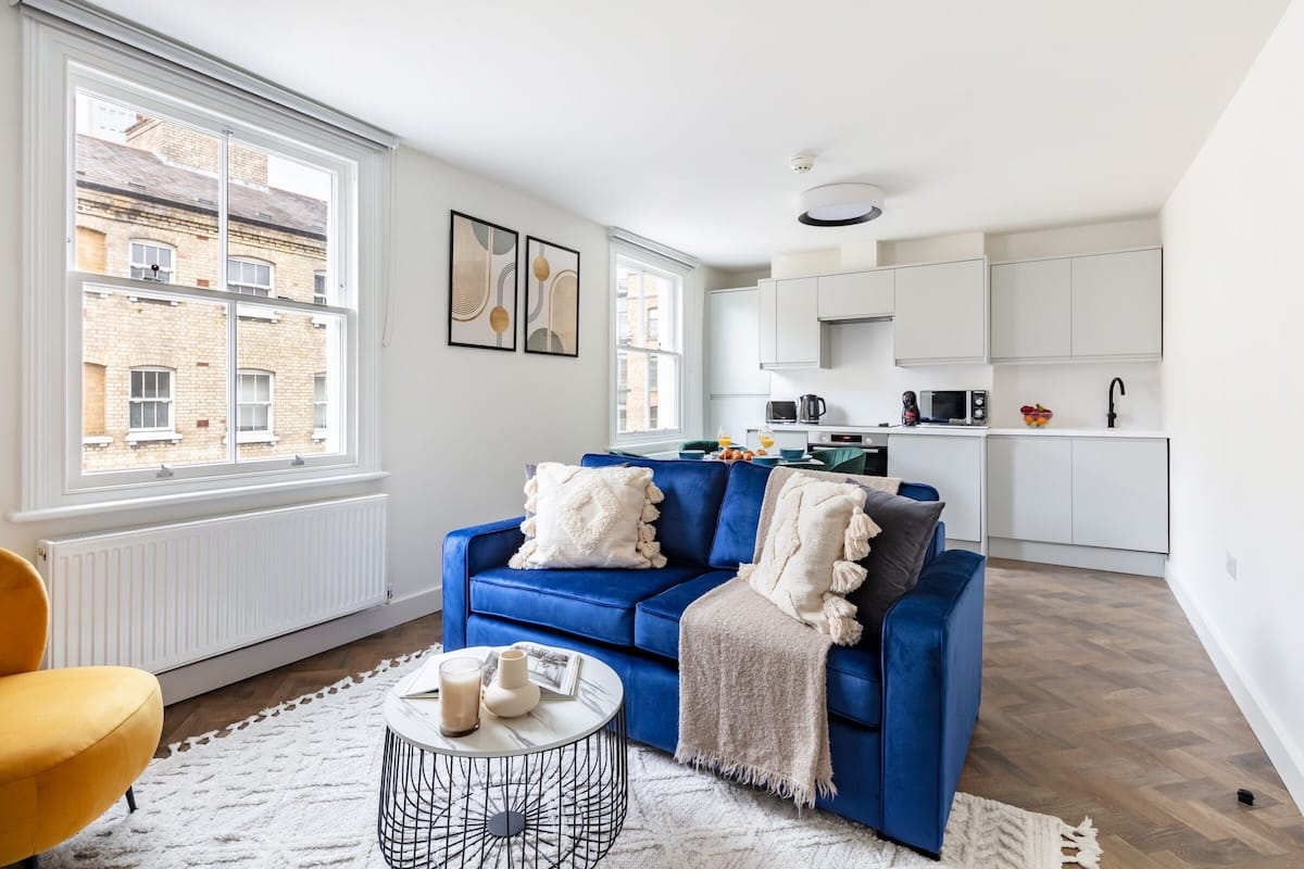Een moderne woonkamer met een blauwe bank, een klein rond salontafeltje en uitzicht op een stadsbeeld door grote ramen.