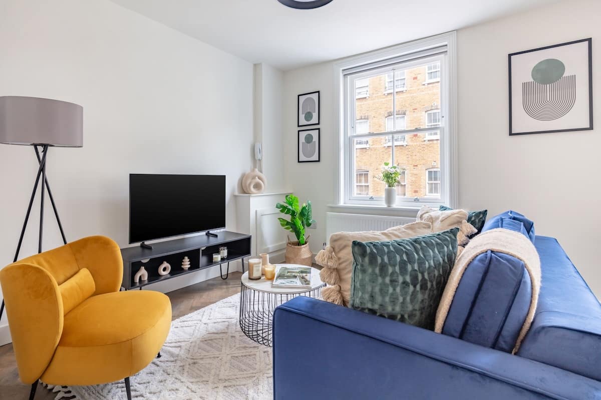 Een moderne woonkamer met een blauwe bank, een mosterdgele fauteuil, een flatscreen televisie, een vloerlamp en een raam met uitzicht op een bakstenen gebouw.