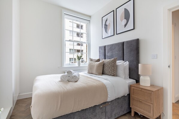 A bedroom with a large bed, a bedside table with a lamp, and a window with a view of buildings. - Modern 1BR Downtown Stay (London)