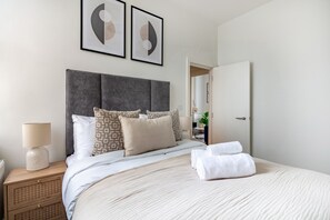 A neatly made bed with a grey headboard, white bedding, and a bedside lamp. There are two framed abstract artworks on the wall above the bed. - Modern 1BR Downtown Stay (London)