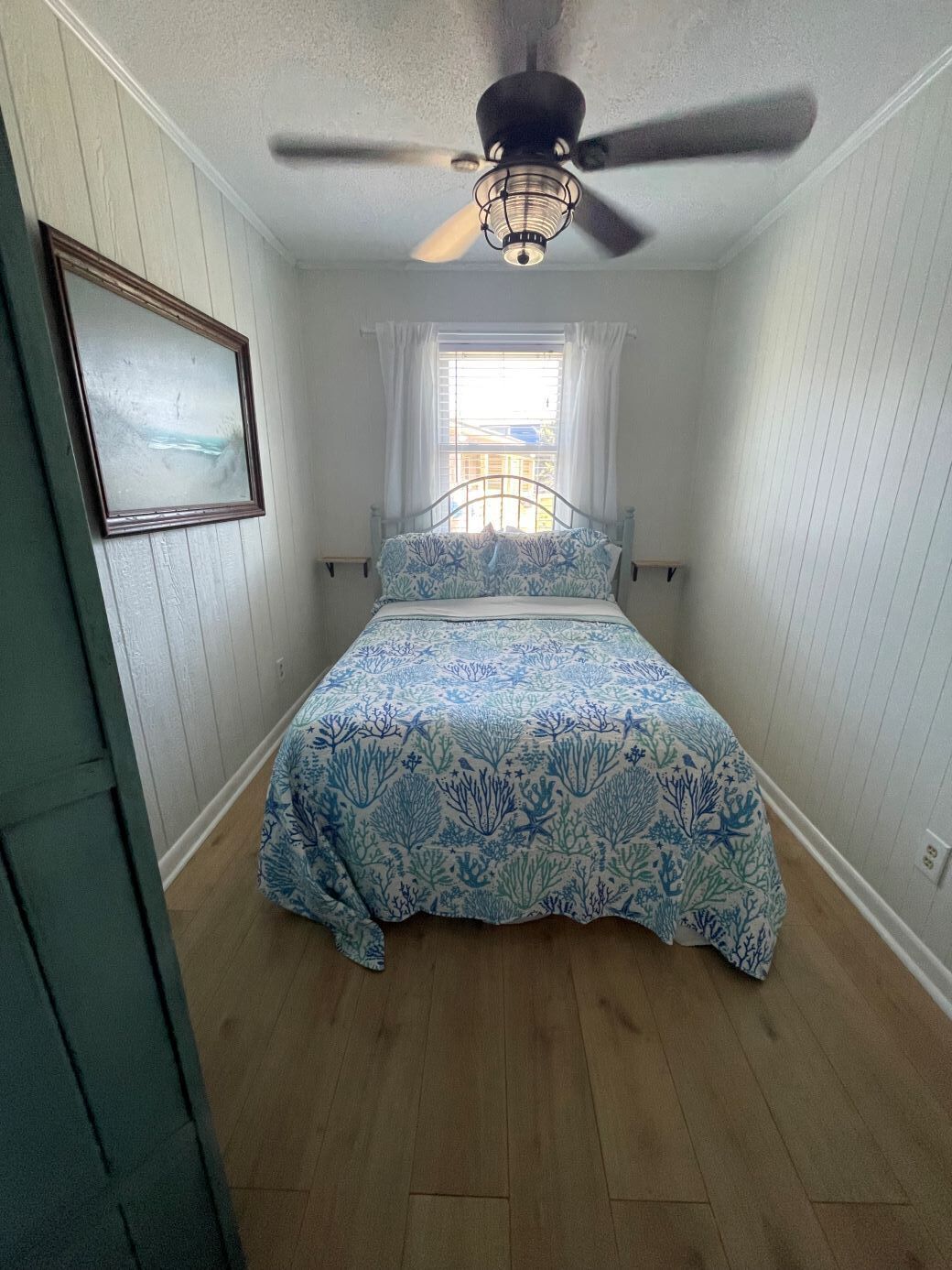 Un dormitorio con cama, ventilador de techo, ventana con cortinas e imagen enmarcada en la pared.