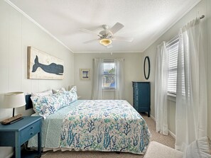 Une chambre à coucher avec un lit, un ventilateur de plafond, une commode, une fenêtre avec des rideaux et un mur orné d’un tableau.