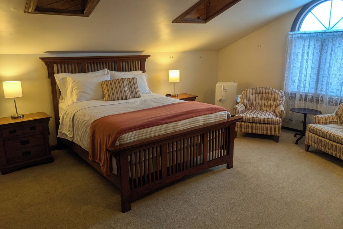 A bedroom with a wooden bed, two bedside tables, a chair, a small table, and a window with curtains.