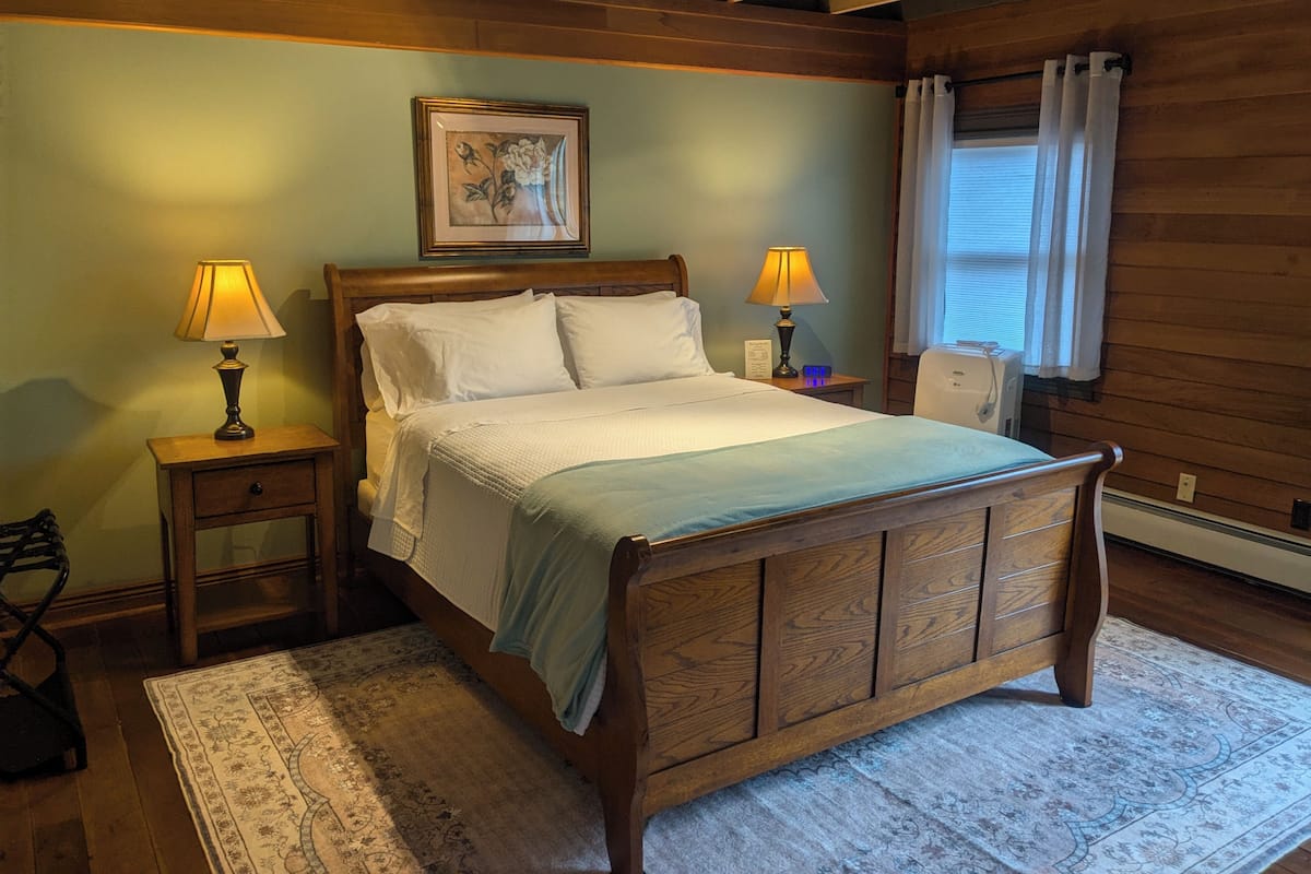 A wooden bed with a headboard, two bedside tables with lamps, a framed picture on the wall, and a window with curtains.