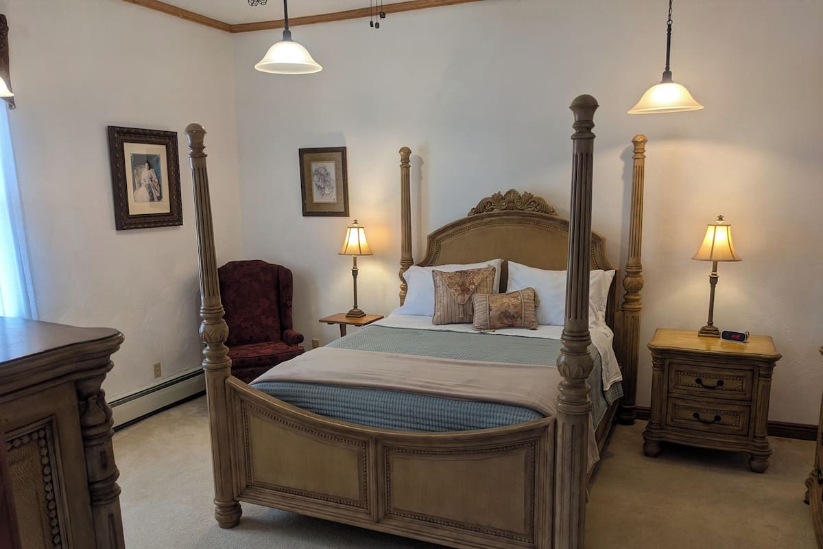 A four-poster bed with a headboard, two bedside tables with lamps, a chair, and framed pictures on the walls.
