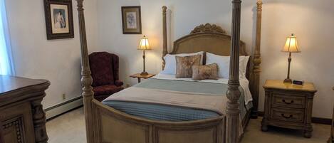 A four-poster bed with a headboard, two bedside tables with lamps, a chair, and framed pictures on the walls.