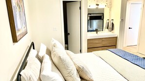 Ein Schlafzimmer mit Bett, Kissen, Nachttisch, Spiegel, Badezimmer mit Waschbecken und einer Tür.