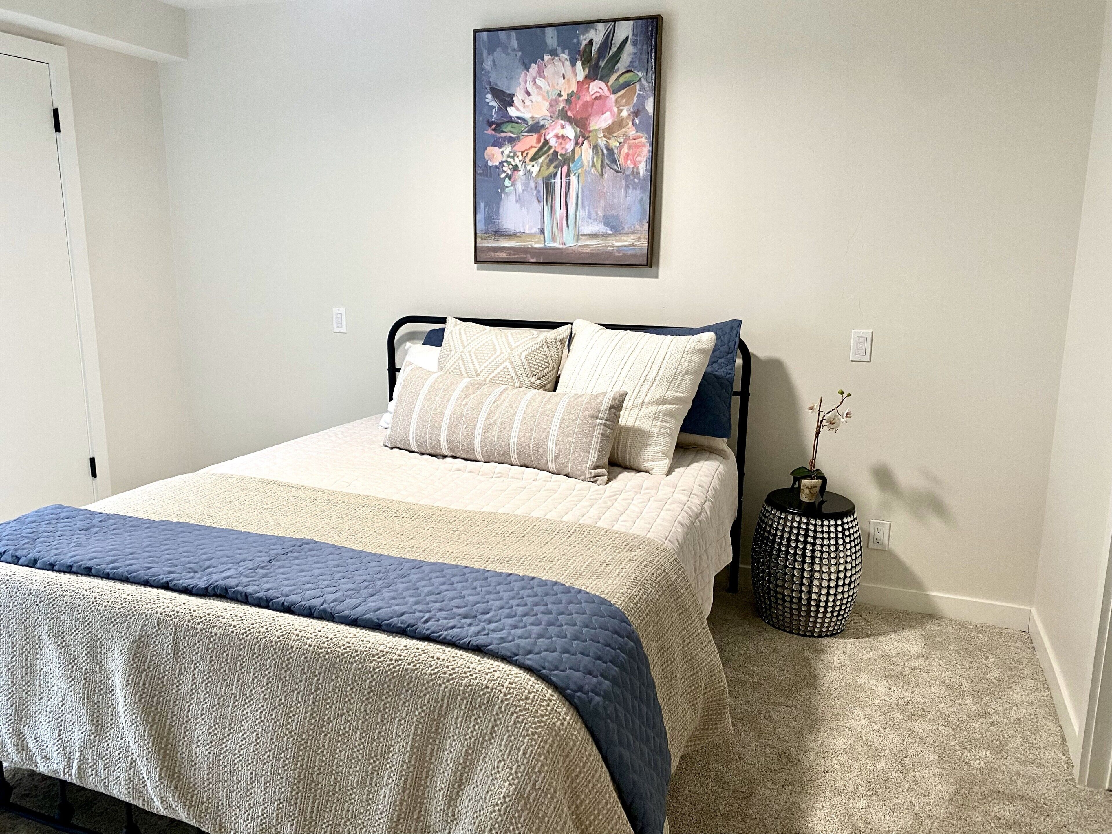A neatly made bed with a quilted cover, a bedside table with a vase, and a painting on the wall.