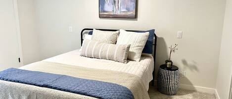 A neatly made bed with a quilted cover, a bedside table with a vase, and a painting on the wall.