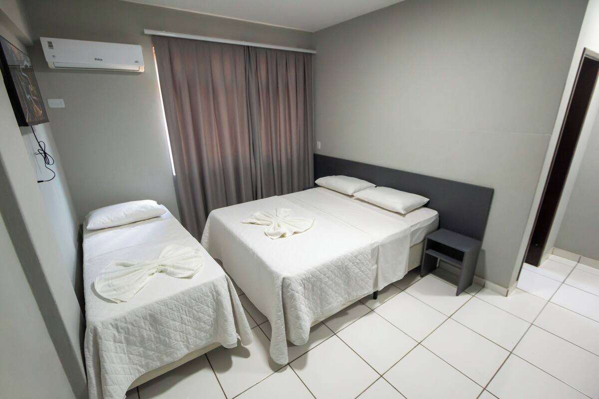 A hotel room with two beds, a small bedside table, a wall-mounted air conditioner, and a window with curtains.