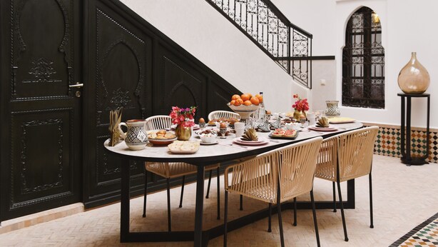Un espace repas avec une longue table dressée pour un repas, entouré de chaises en osier, avec un escalier et des éléments décoratifs en arrière-plan.