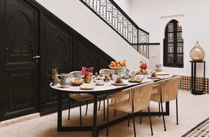 A dining area with a long table set for a meal, surrounded by wicker chairs, with a staircase and decorative elements in the background. - Riad Arraha Retreat Elegance tranquility and pools (Marrakesh)