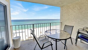 A balcony with a glass table and chairs, overlooking a beach with clear blue water and a sandy shore.