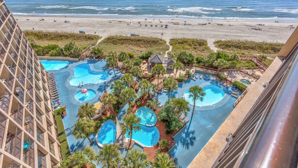 A hotel with multiple swimming pools surrounded by palm trees, overlooking a beach.