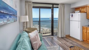 A living room with a sofa, a lamp, a painting, a view of the ocean, and a kitchen area with wooden cabinets.