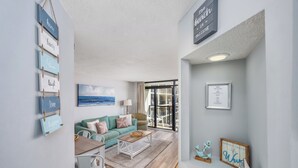 A modern living room with a teal sofa, a coffee table, and a dining area with a white table and chairs. There is a large window with a view of the ocean, and a sign on the wall that reads "The beach is calling."