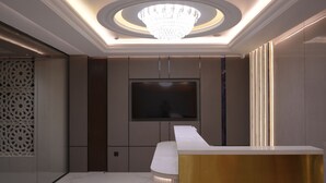 A modern interior with a large TV mounted on the wall, a white reception desk, and a decorative ceiling light. - Raam Leela Hotel (Faizabad)
