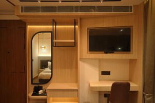 A modern hotel room with a wooden wardrobe, a desk with a chair, a flat-screen TV mounted on the wall, and a mirror reflecting the room.