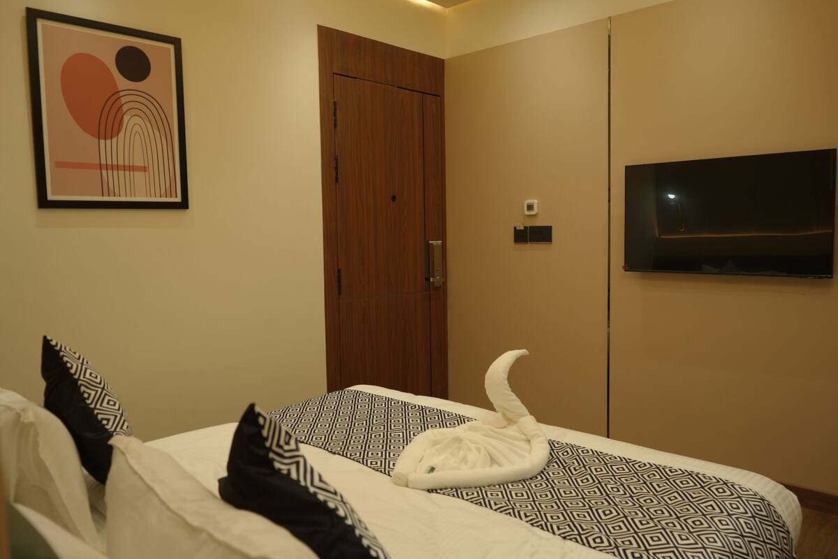 A hotel room with a bed, a framed artwork, a wooden door, a flat-screen TV, and a towel on the bed.