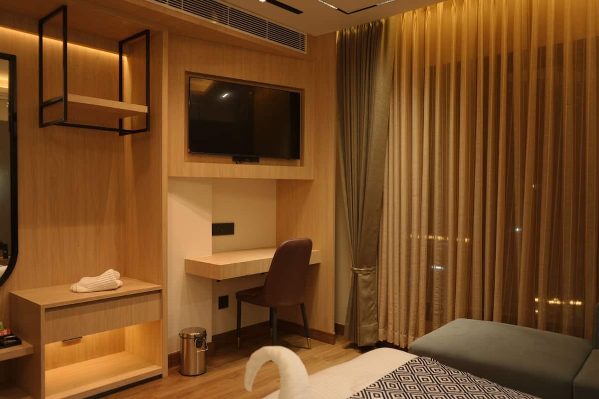 A modern hotel room with a wooden desk, a television mounted above it, a comfortable bed, and a large window with curtains.