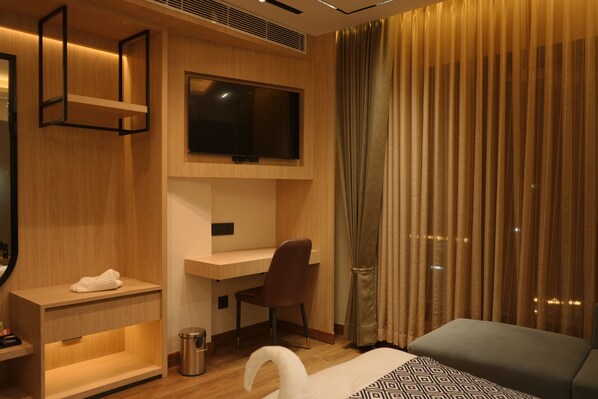 A modern hotel room with a wooden desk, a television mounted above it, a comfortable bed, and a large window with curtains.