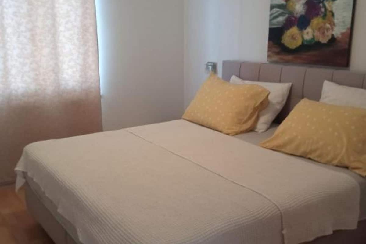 A neatly made bed with a white and beige bedspread, two yellow pillows, and a headboard with a floral painting.