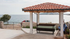 A covered outdoor seating area with a bench, a car parked nearby, and a view of the sea. - Apartments Ruzarija (Marina)