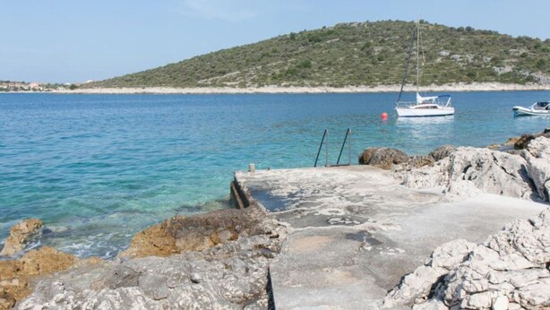 A rocky waterfront with clear blue water, a small boat docked, and a hill in the background. - Apartments Ruzarija (Marina)