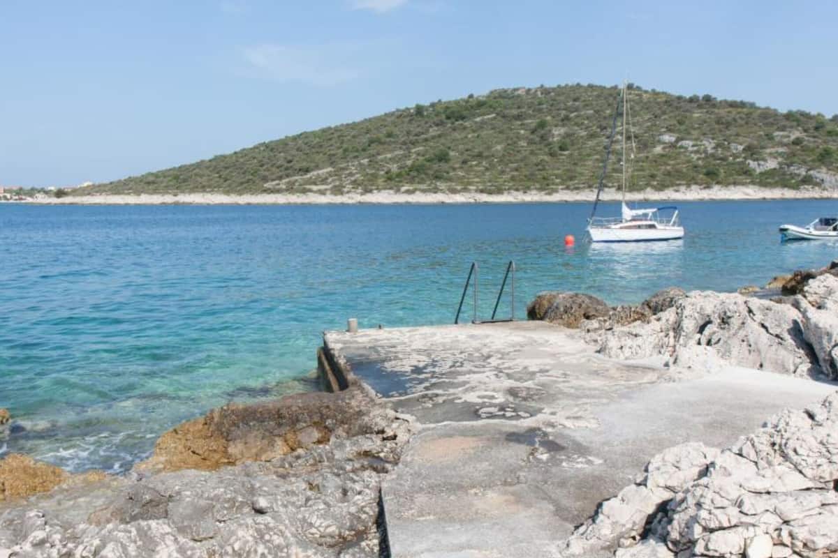 A rocky waterfront with clear blue water, a small boat docked, and a hill in the background.