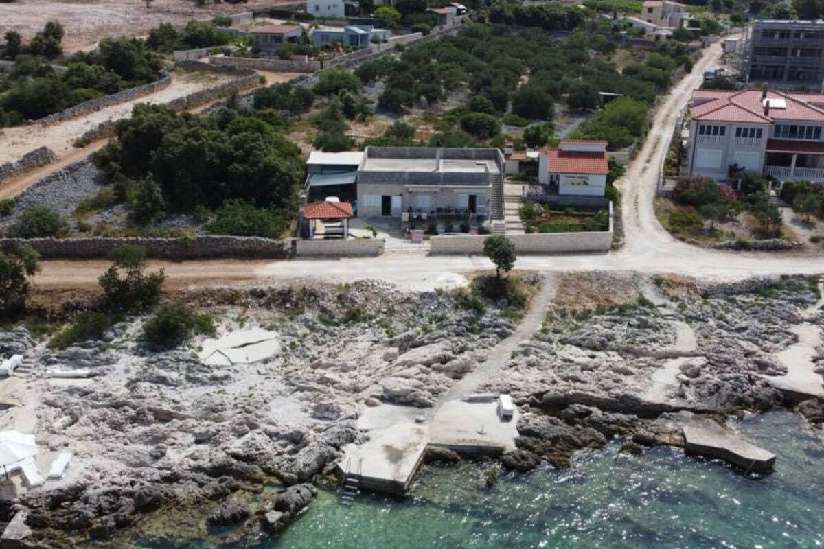 An aerial view of a coastal area with rocky terrain, clear water, and several buildings including a large house with a red roof and a smaller white building.
