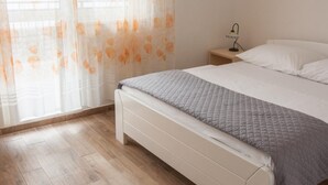 A bedroom with a wooden floor, a bed with a quilted blanket, a nightstand with a lamp, and sheer curtains with a floral pattern. - Apartments Ruzarija (Marina)