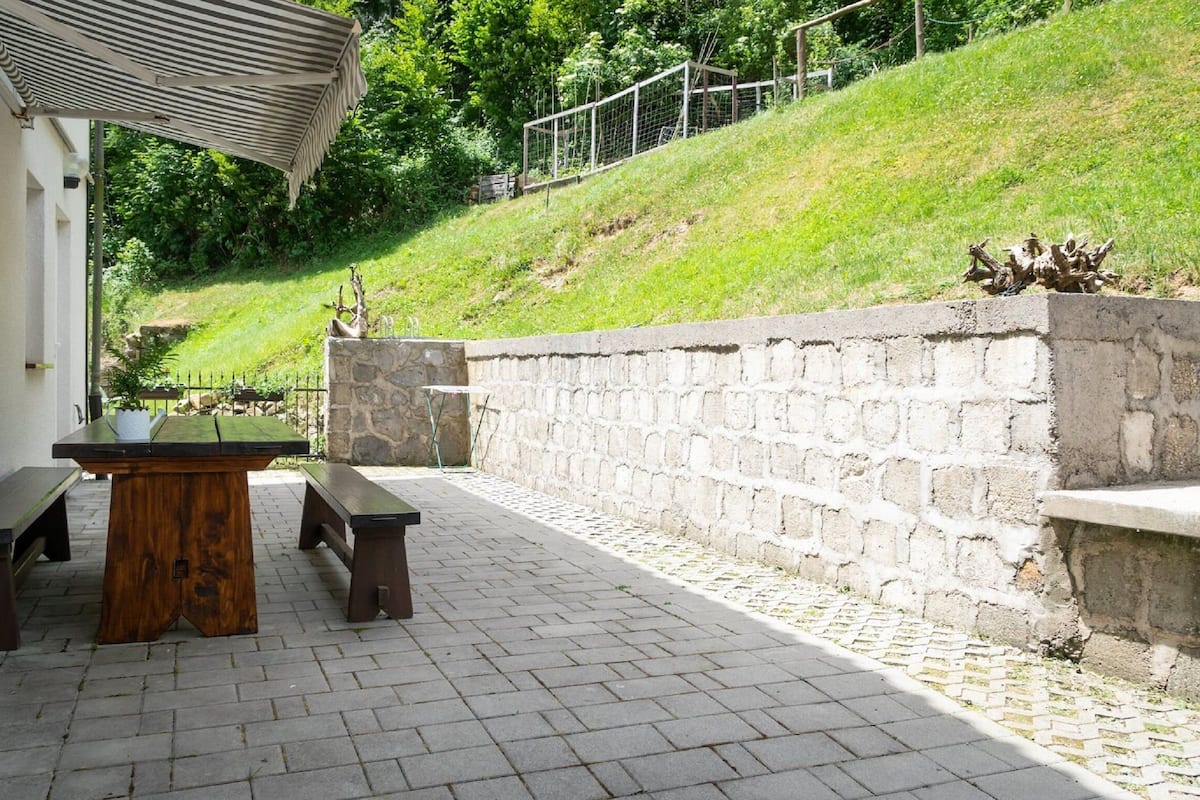 Um pátio externo com mesa e bancos, uma parede de pedra e uma colina gramada ao fundo.