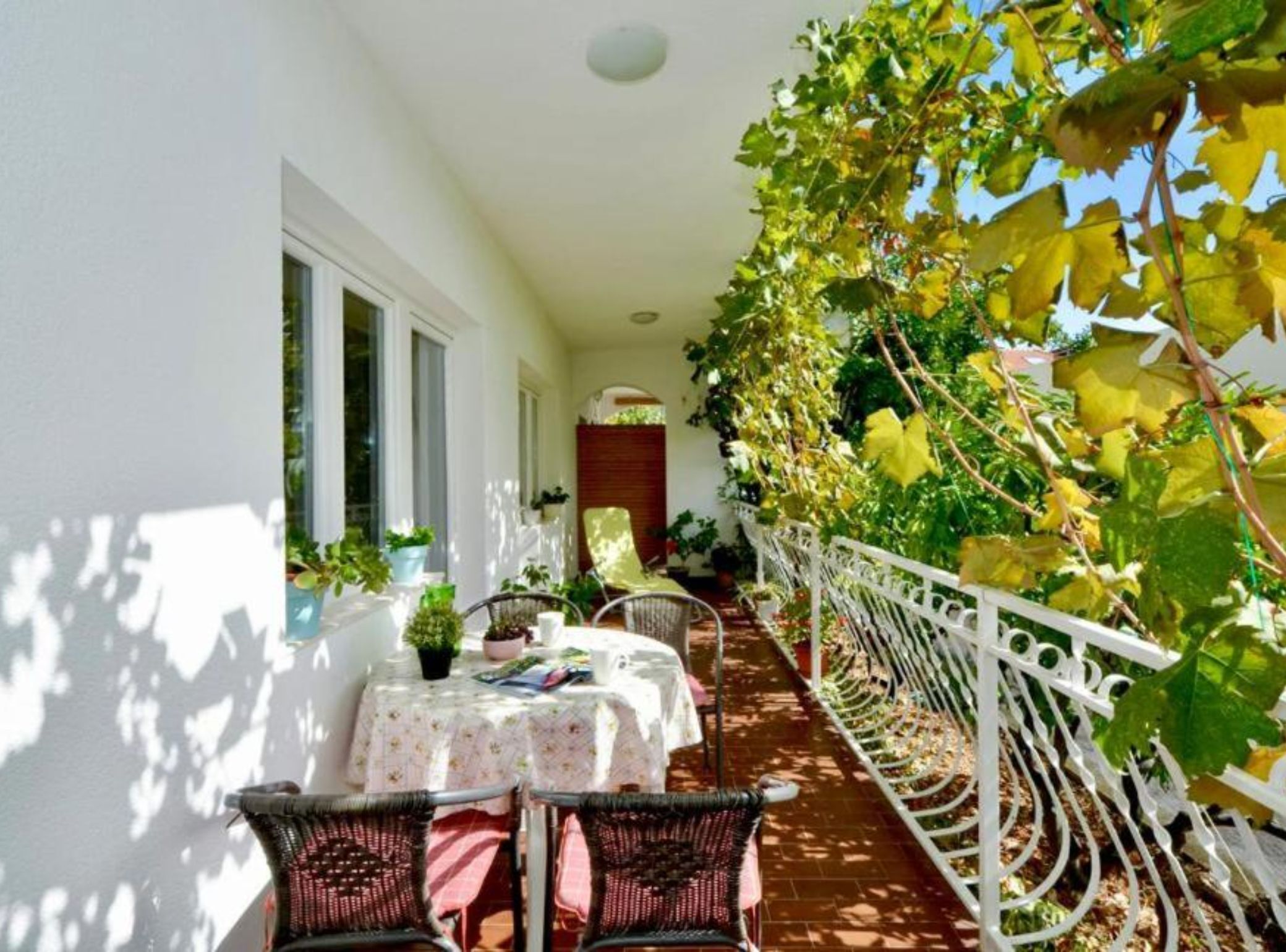A covered outdoor patio with a table and chairs, surrounded by potted plants and a white railing.
