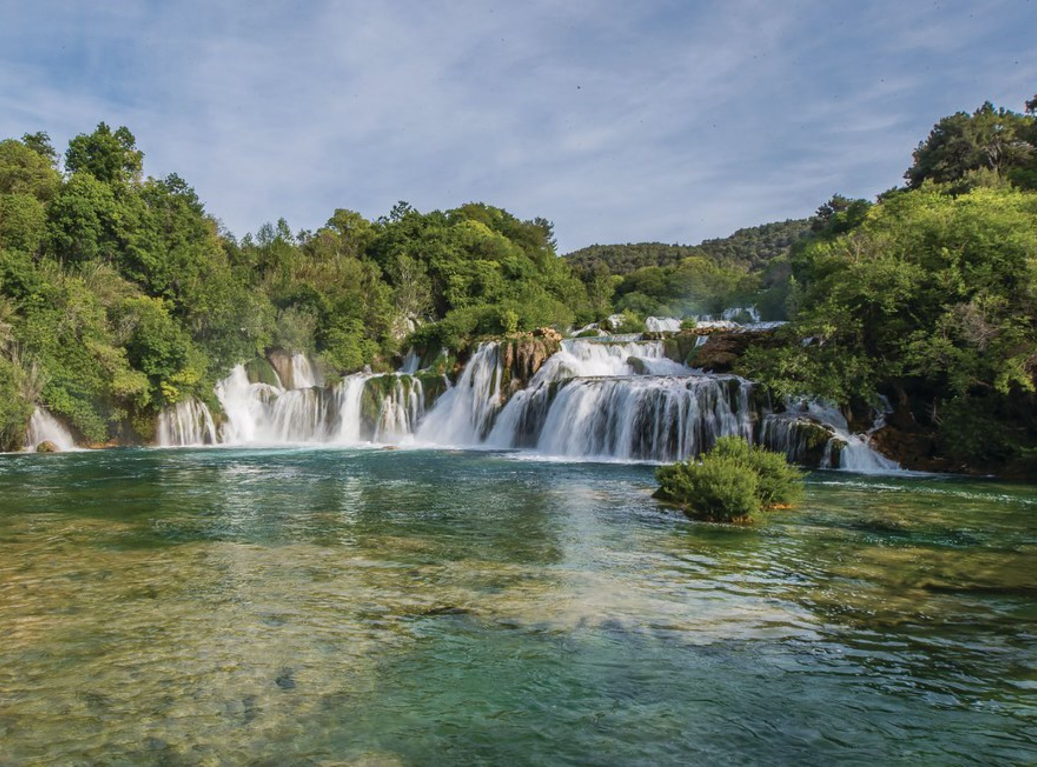 A waterfall cascading into a clear river surrounded by dense green forest.